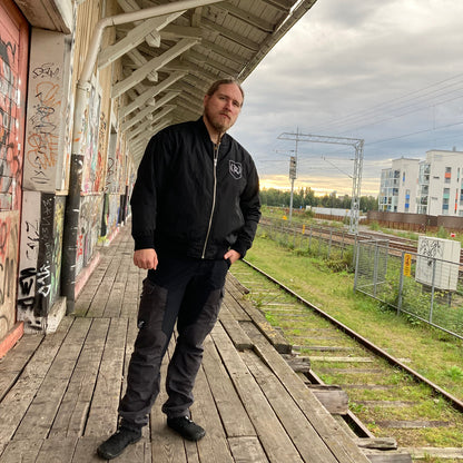 Rurik Fightgear-lentäjänpusakka.
Rurik FG "R" -logo rintapielessä sekä riimulogo selässä monivärisesti brodeerattuna. Kuvassa pilottitakki  Mikan päällä junan laiturilla. ptcumbat.com